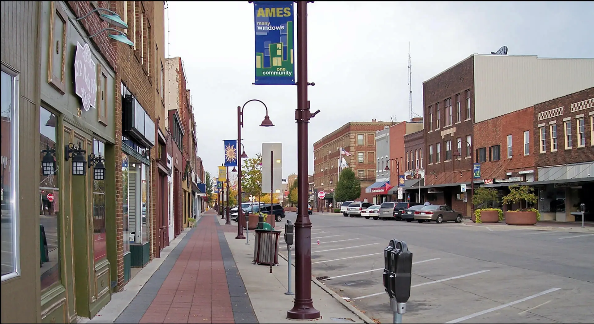 Main Street in downtown Ames, Iowa, with historic brick storefronts, a red brick sidewalk, and a lamppost banner reading Ames, many windows, one community