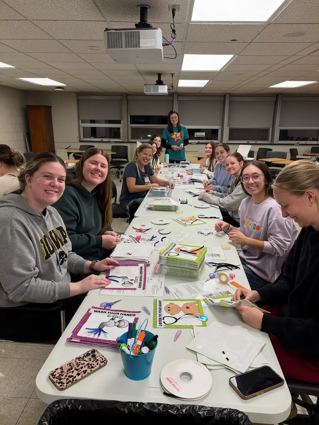 Around a dozen Love From Levi volunteers seated at a long table assembling children's books, with finished Wash Your Hands and Listen to Your Heart pages, ribbon, and markers spread in front of them.
